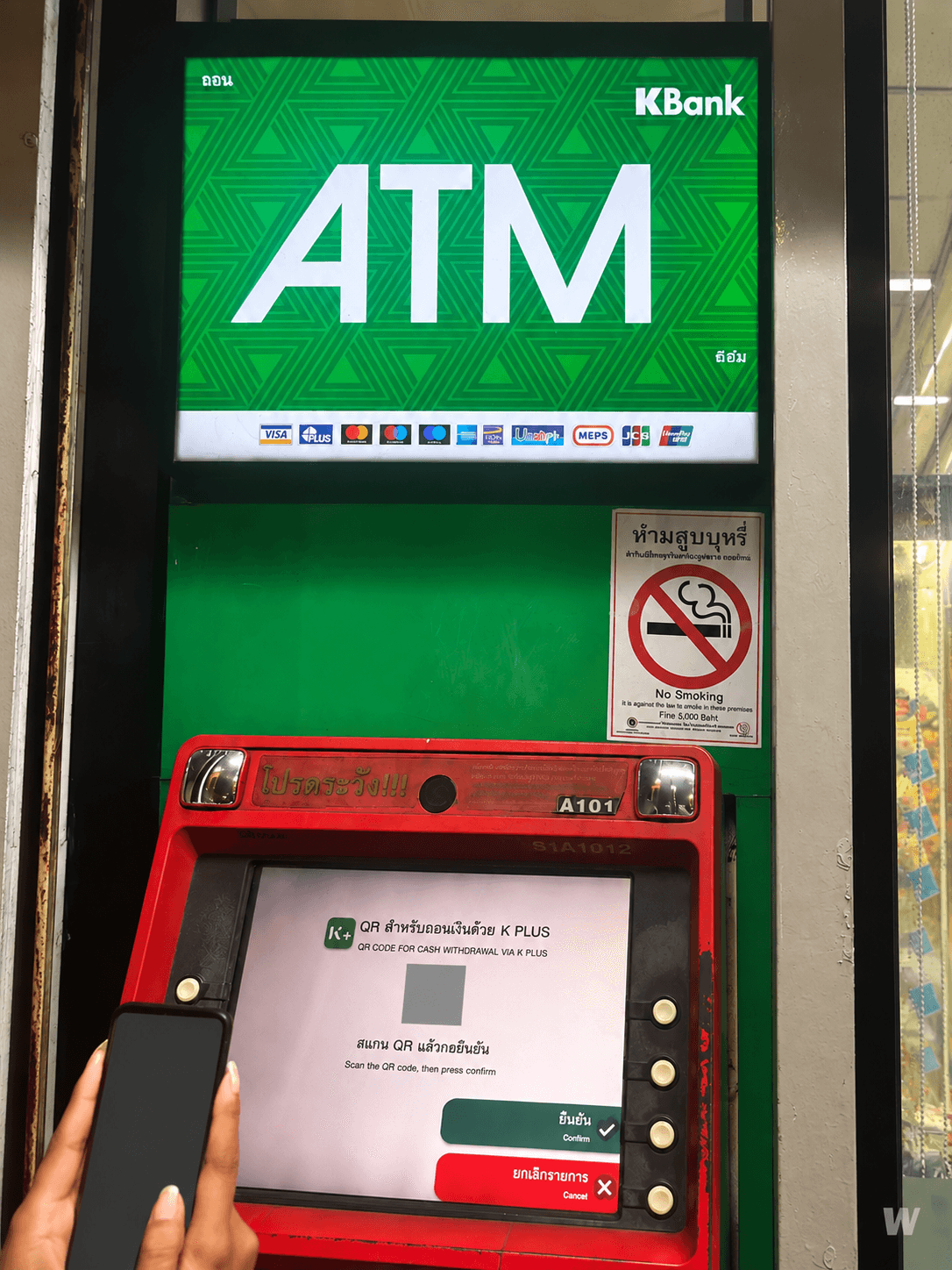 Банкомат KBank с зелёной вывеской; QR-код операции и экран телефона закрыты